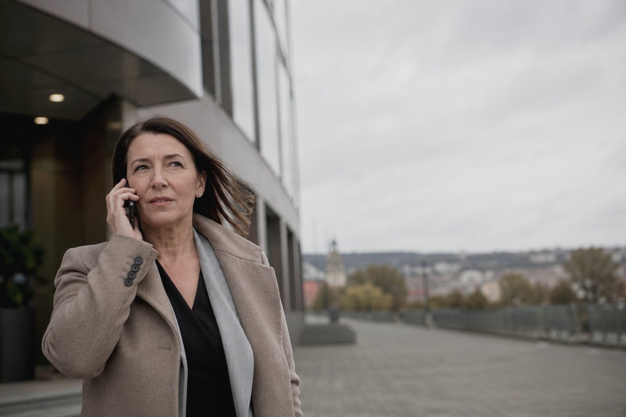 Podnikateľka v béžovom kabáte a sivej šále telefonuje pred modernou pražskou kancelárskou budovou; v pozadí sa pod zamračenou jesennou oblohou rozprestiera panoráma pražských striech s vežou kostola.