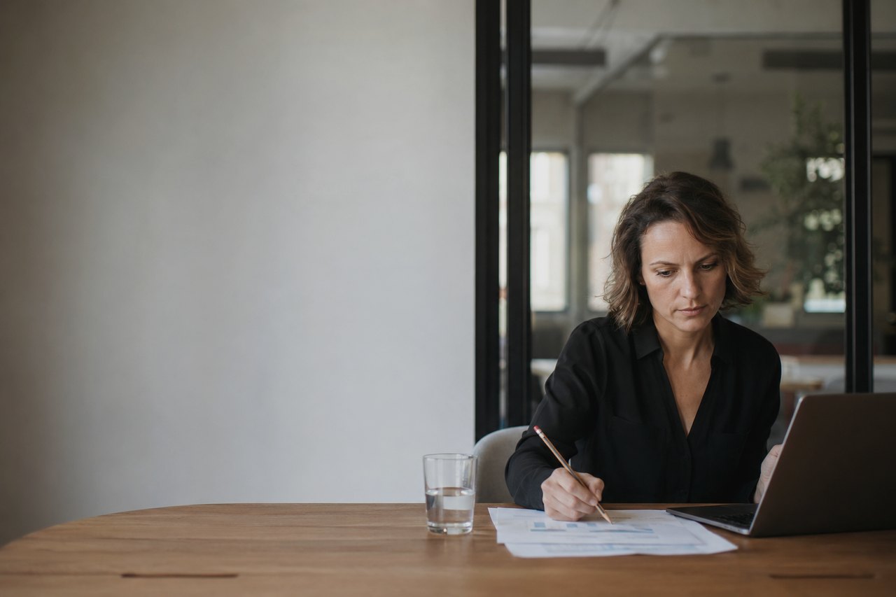 Žena v čiernej košeli sedí pri drevenom stole, ceruzkou si prechádza vytlačené grafy a tabuľky, vedľa má otvorený notebook a pohár vody; v pozadí presklená stena a svetlá moderná kancelária.